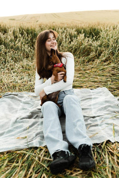 Linda garota segurando um cachorro bassê nos braços. menina brincando com um cachorro em um campo de trigo ao pôr do sol. retrato de uma menina com um animal de estimação favorito