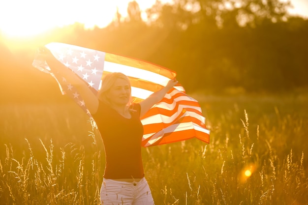 Linda garota segurando uma bandeira americana ao vento em um campo de centeio. Paisagem de verão contra o céu azul. Orientação horizontal.