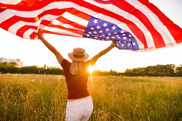 Linda garota segurando uma bandeira americana ao vento em um campo de centeio. Paisagem de verão contra o céu azul. Orientação horizontal.