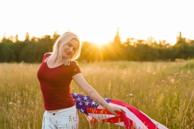 Linda garota segurando uma bandeira americana ao vento em um campo de centeio. Paisagem de verão contra o céu azul. Orientação horizontal.