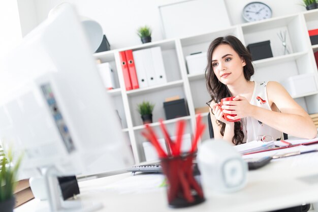 Linda garota senta-se na mesa de escritório, olha para a tela do computador e tem uma caneca vermelha nas mãos.