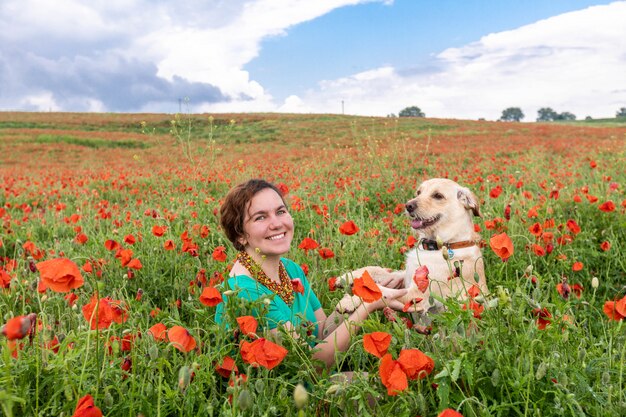 Foto linda garota senta-se no meio de um campo de papoulas e olha para o cachorro