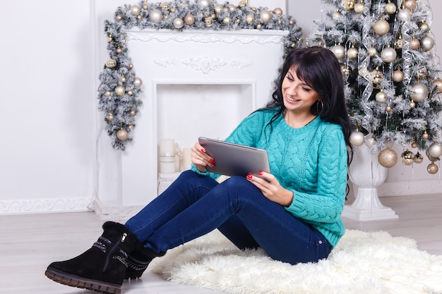Linda garota sentada dentro de casa na decoração de Natal, segurando um tablet pc branco.