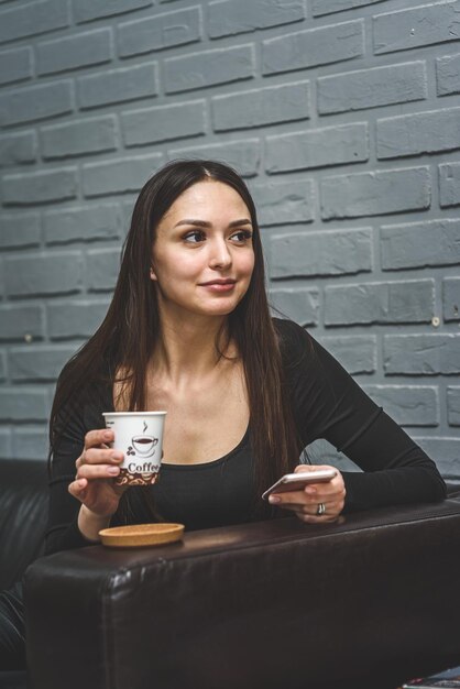 Foto linda garota sentada em um sofá de couro no salão tomando café e falando ao telefone