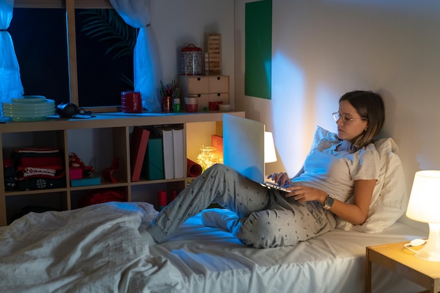 Linda garota sentada na cama à noite, trabalhando com seu laptop no meio da noite