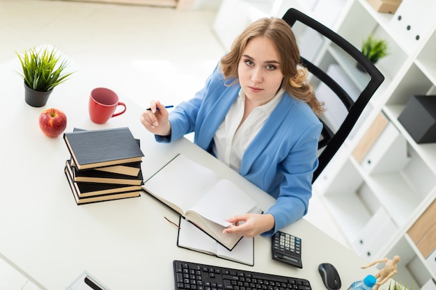 Foto linda garota sentada na mesa no escritório, segurando uma caneta na mão e lendo um livro.