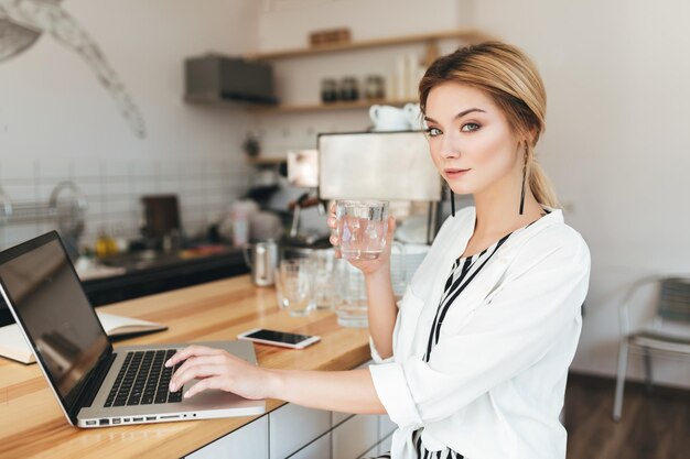 Linda garota sentada no balcão com um copo de água na mão e olhando pensativamente na câmera no café Moça bonita com cabelo loiro trabalhando em seu laptop na cafeteria