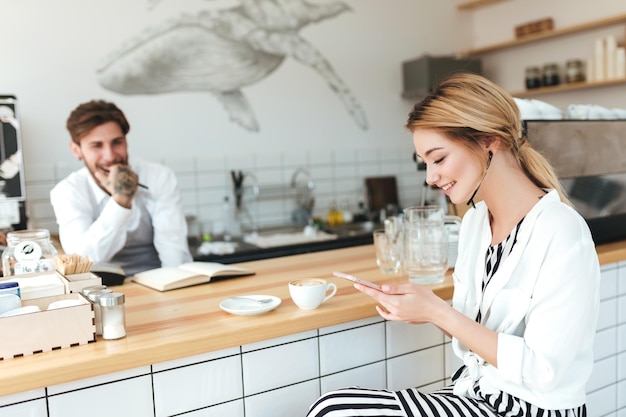 Linda garota sentada no balcão com uma xícara de café e olhando alegremente em seu celular enquanto barista no fundo olhando para ela Linda garota com cabelo loiro tomando café na cafeteria