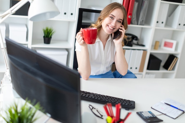 Foto linda garota sentada no escritório, segurando uma caneca na mão e falando ao telefone.