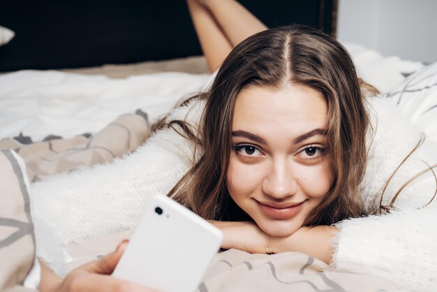 Linda garota sonolenta de pijama branco deitada na cama à noite, quer dormir, sorrindo