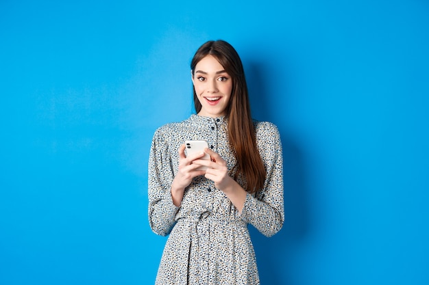 Linda garota sorridente com cabelo longo natural, usando vestido, usando smartphone e parecendo feliz, de pé contra o azul.