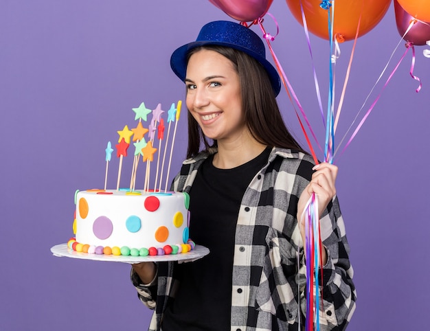 Linda garota sorridente com chapéu de festa segurando balões com bolo isolado na parede azul