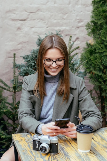 Linda garota sorridente com óculos, sentada em um café, segurando o smartphone nas mãos e conversando