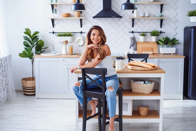 Linda garota sorridente em t-shirt branca senta-se na elegante cozinha branca de manhã.