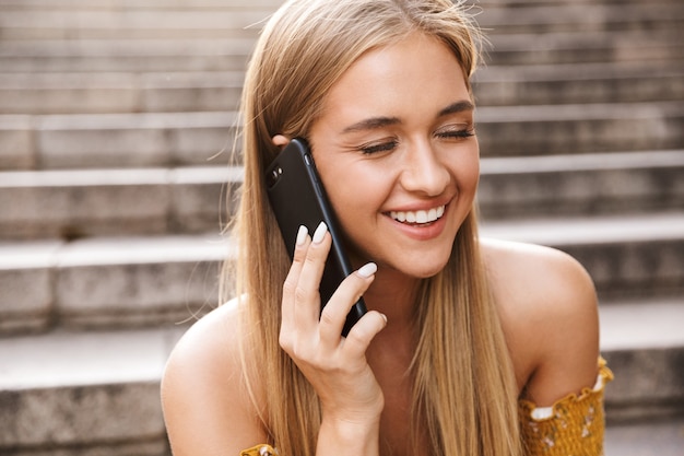 Linda garota sorridente falando no celular enquanto está sentada na escada ao ar livre