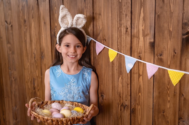 Linda garota sorridente fica perto da parede de madeira e segura uma cesta com ovos coloridos.