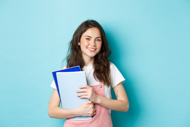 Linda garota sorridente segurando cadernos e olhando alegre para a câmera, estudando na faculdade ou universidade, em pé sobre um fundo azul