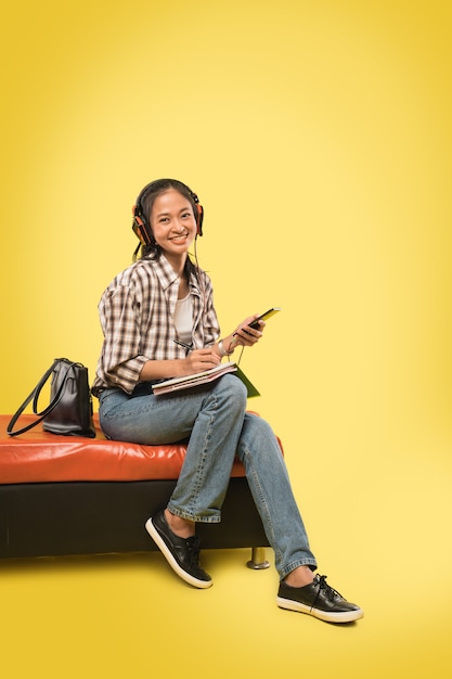 Linda garota sorridente usando fones de ouvido e escrevendo usando uma caneta no livro enquanto segura o celular