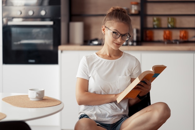 Linda garota tomando café e lendo um livro na mesa da cozinha em casa