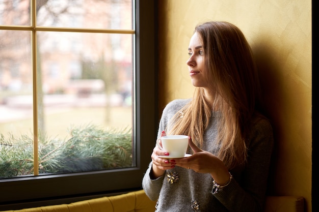 Linda garota tomando café ou chá no café e olhando pela janela.