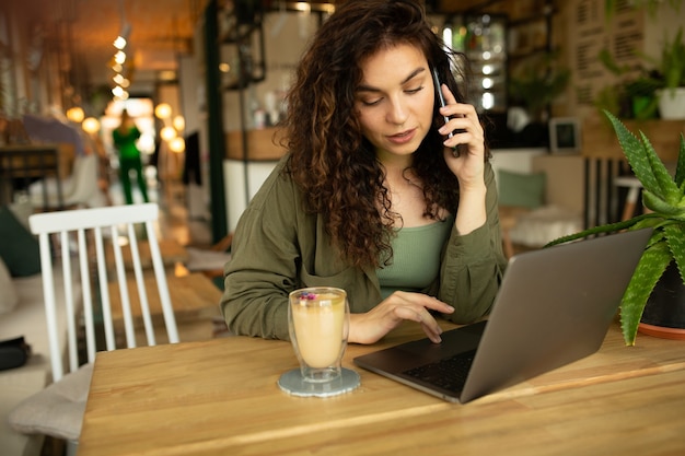Linda garota trabalhando em uma cafeteria com uma freelancer mulher laptop segurando um telefone conectado à internet através do computador, fazendo compras online