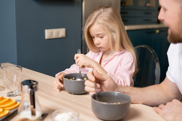 Linda garota triste colocando a colher na tigela com muesli enquanto toma café da manhã na mesa na cozinha com o pai dela sentado perto