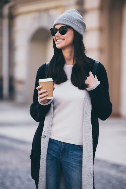 Linda garota urbana. Mulher jovem e bonita em óculos de sol segurando uma xícara de café e olhando para longe enquanto caminha ao ar livre