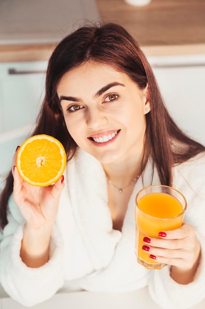 Linda garota usa roupão branco com copo de suco de laranja e cozinha de frutas vista do pôr do sol da manhã