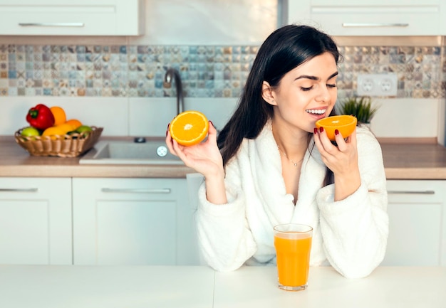 Linda garota usa roupão branco com copo de suco de laranja e cozinha de frutas vista do pôr do sol da manhã