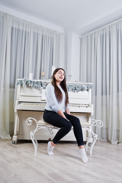 Linda garota usar suéteres quentes e calças pretas contra o velho piano com decoração de Natal no quarto branco.