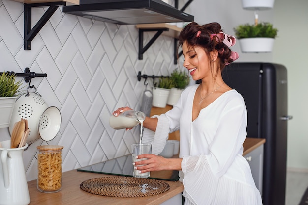 Linda garota vestida com roupão branco derrama o leite de uma jarra em um copo, na elegante cozinha aconchegante. Conceito de alimentação e dieta saudável.