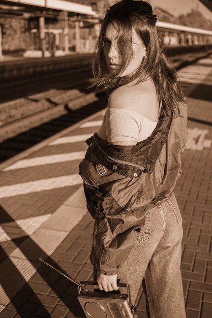 Linda garota vestida no estilo dos anos 90 com receptor de rádio portátil nas mãos posando na plataforma da estação de trem