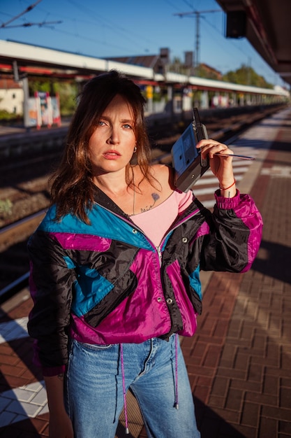 Linda garota vestida no estilo dos anos 90 com receptor de rádio portátil nas mãos posando na plataforma da estação de trem