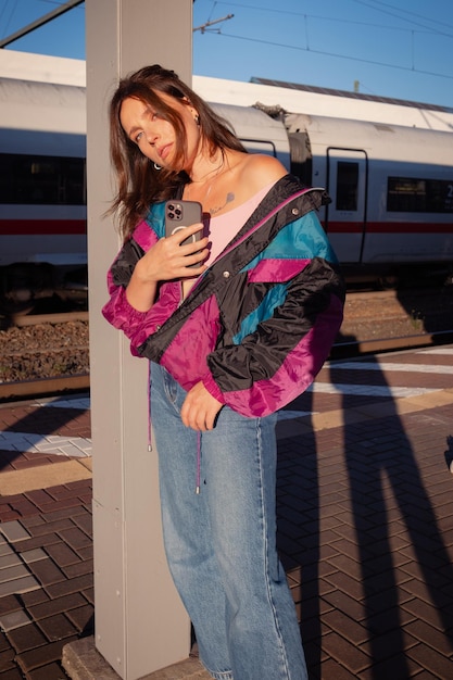 Linda garota vestida no estilo dos anos 90 com smartphone moderno nas mãos posando na plataforma da estação de trem