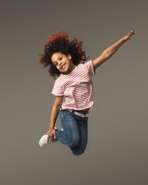 Foto linda garotinha afro-americana pulando um fundo cinza do estúdio, copie o espaço. foto de estúdio de uma criança feliz e fofa