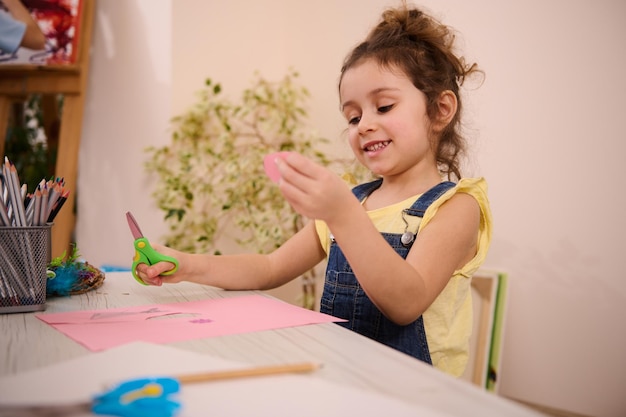 Linda garotinha caucasiana senta-se em uma mesa e sorri olhando para a câmera enquanto desenha e corta papel colorido durante a aula de arte Educação infantil e conceito de entretenimento