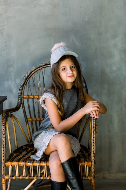 Linda garotinha com cabelos castanhos compridos e olhos azuis, sentado em uma poltrona vestida de vestido cinza e um chapéu