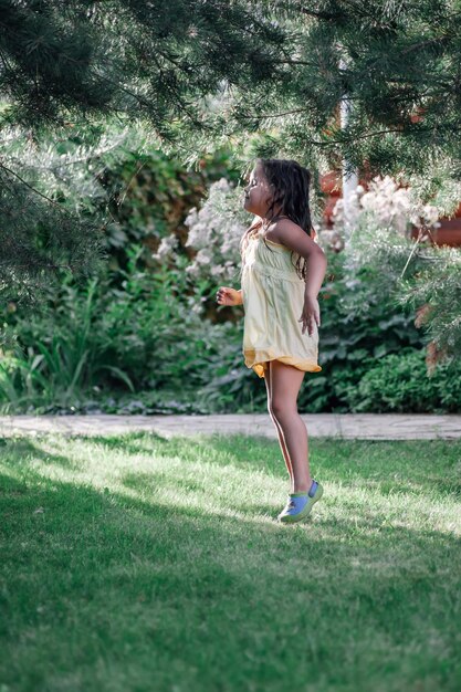 Linda garotinha com os olhos fechados se divertindo e prestes a pular em pé sob galhos de pinheiro usando vestido leve de verão e chinelos no parque durante o dia Fundo incrível cheio de verde