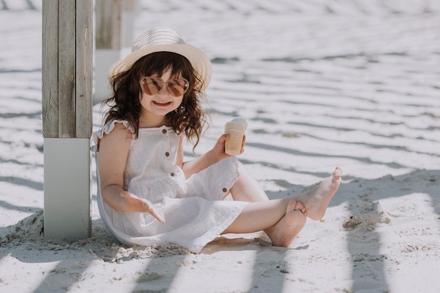 Linda garotinha de óculos de sol vestido branco e chapéu comendo sorvete na praia no verão