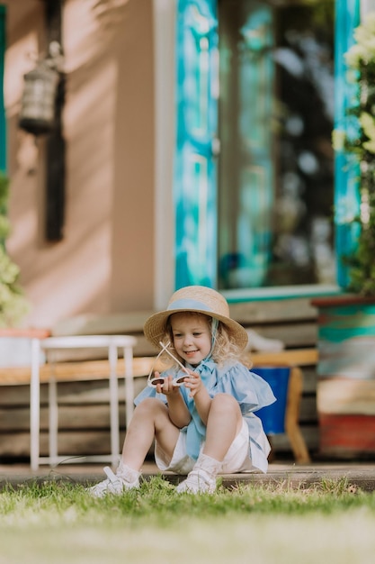 linda garotinha de vestido azul, chapéu de palha e óculos de sol jogando ao ar livre, fundo azul, cartão