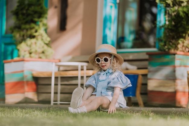 Linda garotinha de vestido azul, chapéu de palha e óculos de sol jogando ao ar livre, fundo azul, cartão