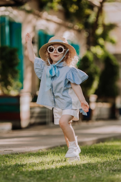 linda garotinha de vestido azul, chapéu de palha e óculos de sol jogando ao ar livre, fundo azul, cartão