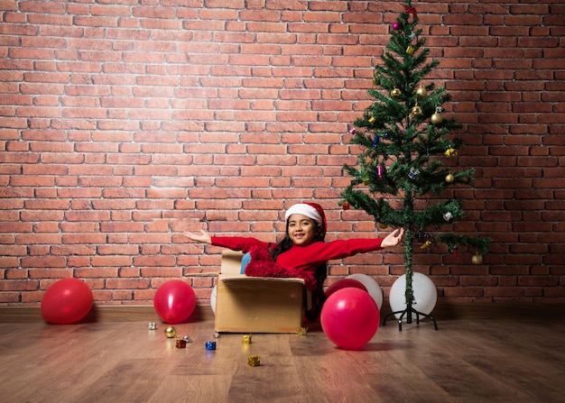 Linda garotinha indiana asiática comemorando o natal enquanto está sentada em uma caixa de papelão contra a parede de tijolo vermelho, interior