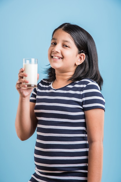 Linda garotinha indiana ou asiática brincalhona segurando ou bebendo um copo cheio de leite, isolado sobre um fundo colorido