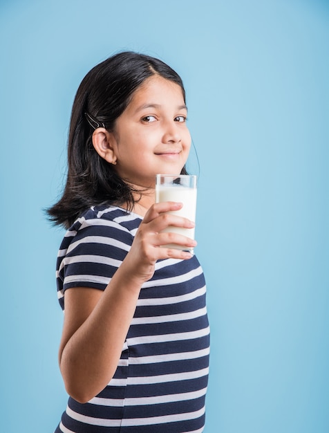 Linda garotinha indiana ou asiática brincalhona segurando ou bebendo um copo cheio de leite, isolado sobre um fundo colorido