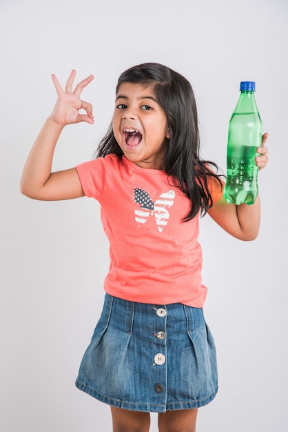 Linda garotinha indiana ou asiática com uma garrafa de estimação de laranja ou bebida gelada de manga ou suco de fruta, bebendo ou segurando em pé isolado sobre um fundo azul ou branco.