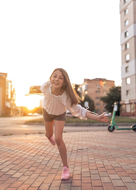 Linda garotinha na cidade ao pôr do sol uma garota de blusa branca e com um chapéu de palha está se divertindo fora de recreação