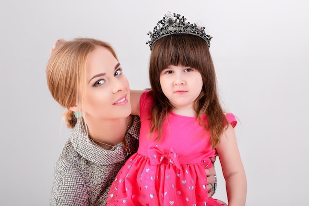 Foto linda garotinha no vestido rosa princesa com uma coroa e linda mãe abraçando em um fundo cinza.