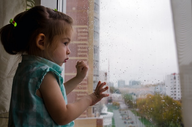 Linda garotinha olhando pela janela na chuva