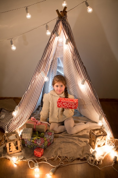 Linda garotinha sentada com um presente nas mãos. Instale a caixa de presente de Natal no feriado à noite na bela sala interior. Ano Novo. Estúdio.